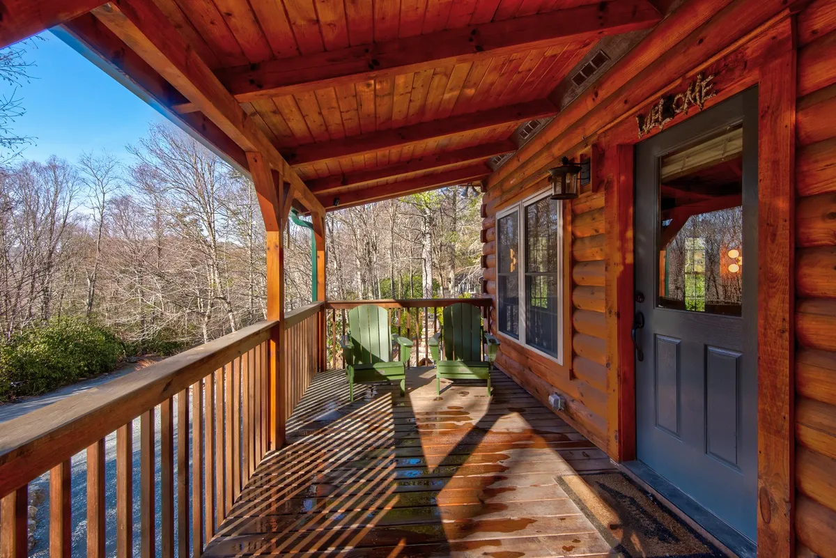 A spacious wooden deck connected to a log cabin, furnished with green chairs and enveloped by trees, offers a tranquil spot to unwind. The deck overlooks serene natural surroundings, ideal for enjoying the fresh mountain air or a peaceful morning.