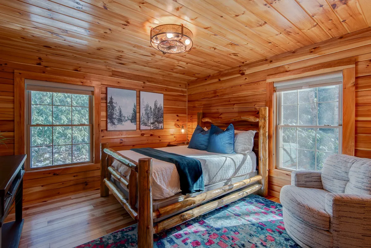 A welcoming bedroom with rustic log bed, large windows letting in abundant natural light, an armchair, and wall artwork. The space evokes the classic log cabin feel with plenty of room to relax and unwind.