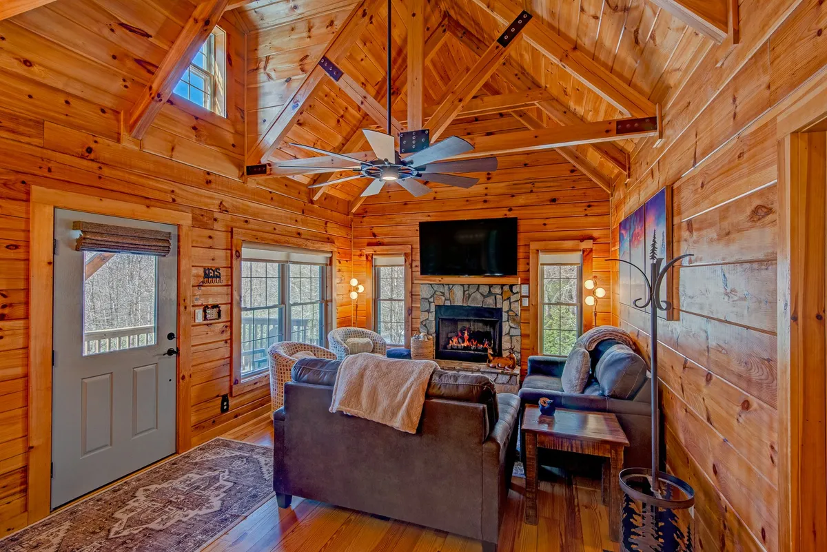 A spacious, inviting den with a grand stone fireplace as the focal point, complemented by a wooden ceiling, large windows streaming in natural light, and a comfortable seating area arranged for both relaxation and conversation. The warm, rustic decor creates a cozy mountain retreat atmosphere.