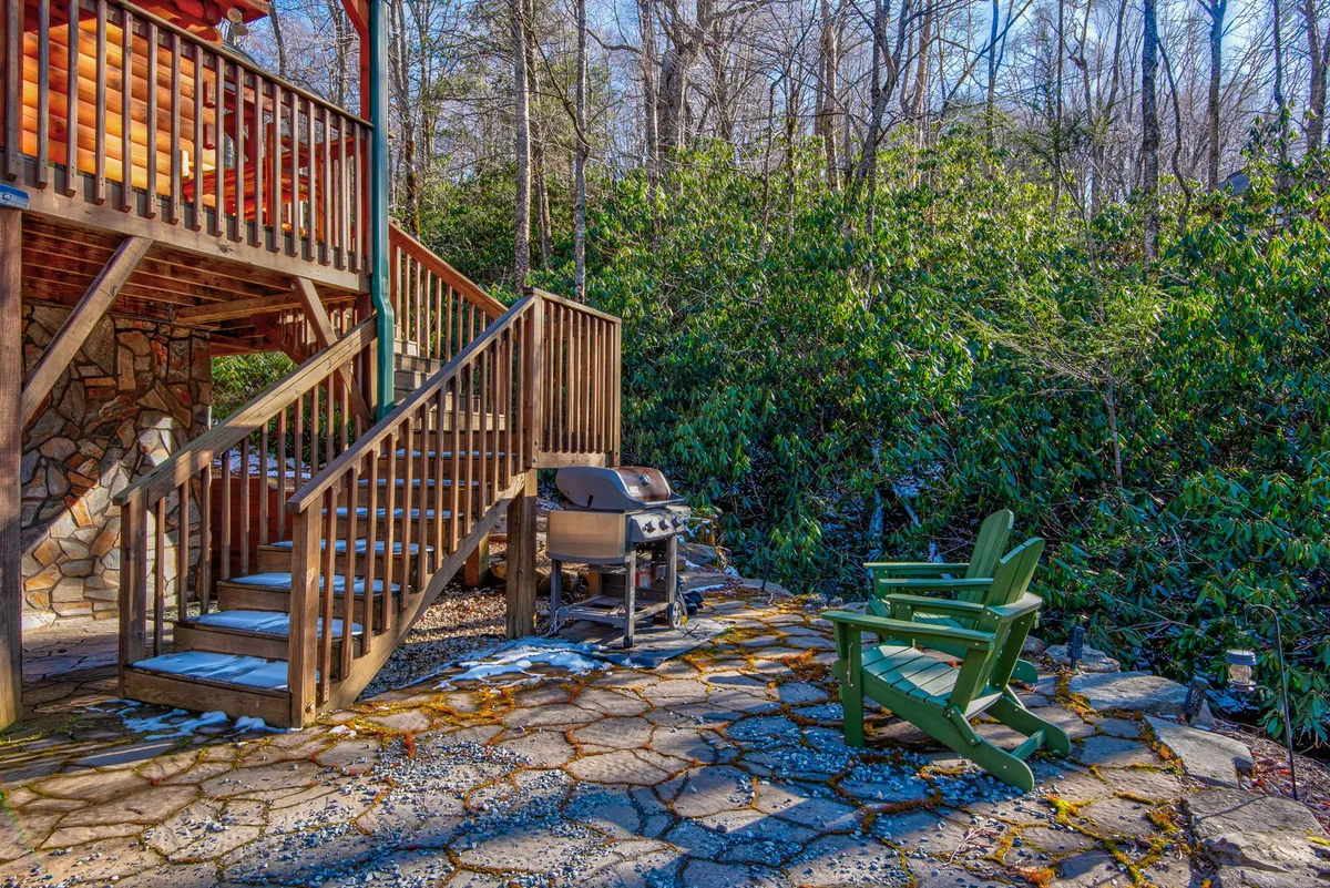 An inviting stone patio features green Adirondack chairs arranged for conversation, a sturdy wooden staircase leading down from the house, and a barbecue grill perfect for outdoor cooking. The setting is surrounded by lush greenery, emphasizing the connection to nature and providing a comfortable place for groups to gather after an adventurous day.