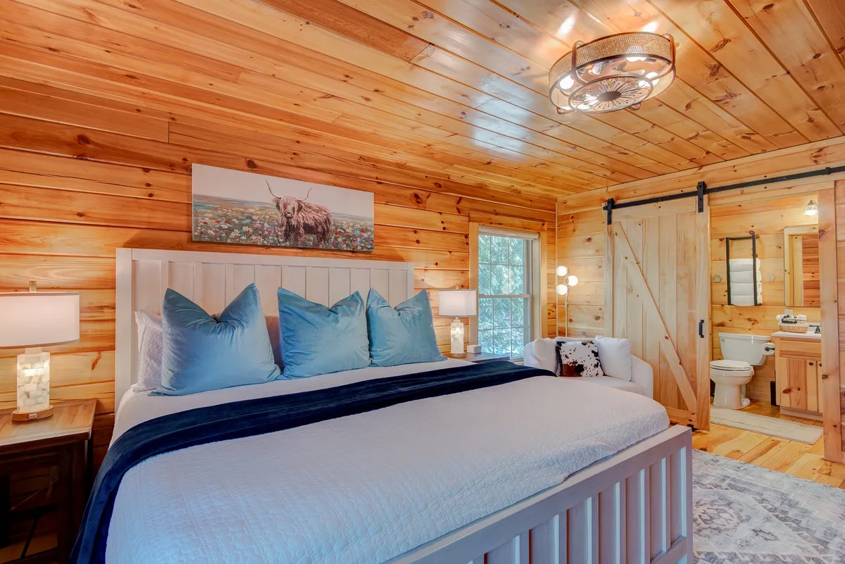 A serene bedroom featuring a plush bed with soft pillows, wooden wall paneling, decorative artwork, and a comfortable seating area. Soft morning light spills through the window, creating a cozy and inviting atmosphere.