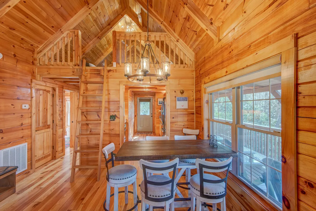 A welcoming dining area with a large wooden table under a stylish chandelier, flanked by expansive windows offering views to the outdoors. A staircase leads up to a loft, hinting at ample space for group activities.
