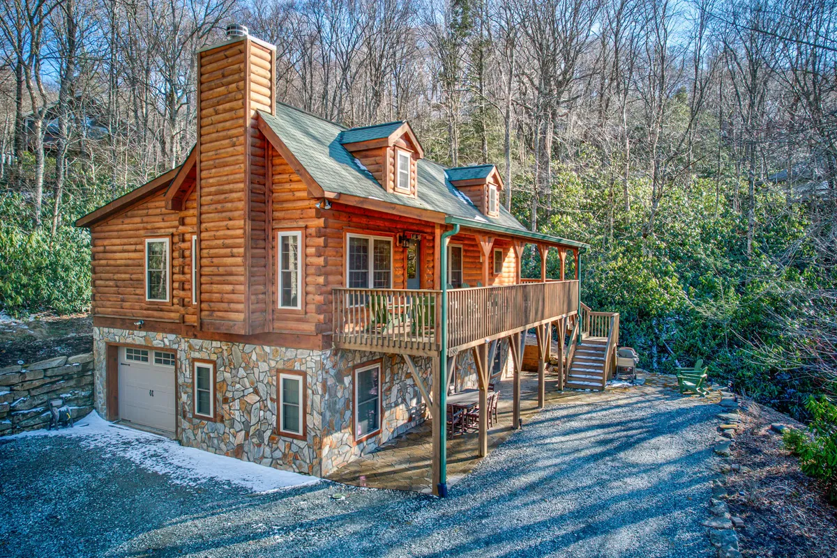 This exterior image showcases the log cabin's inviting deck and stone foundation, surrounded by natural greenery and trees. The deck is set up for gatherings, with space to relax and enjoy the mountain air, perfectly illustrating the cabin's proximity to outdoor adventures.