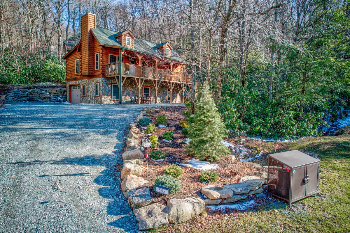 The cozy log cabin exterior features a welcoming front porch and landscaped garden, set against a backdrop of trees and a gravel driveway. The porch invites relaxation and hints at the comfort within, while the well-maintained landscaping underlines care and attention to the property.