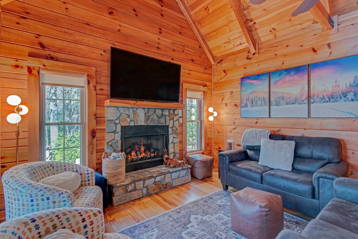 A cozy living area featuring a fireplace as the focal point, surrounded by a plush sofa and armchairs. Large windows offer views of the outdoors, adding a sense of openness and connection to nature. Artwork decorates the wall, and the intimate setting is ideal for unwinding in peace and comfort.