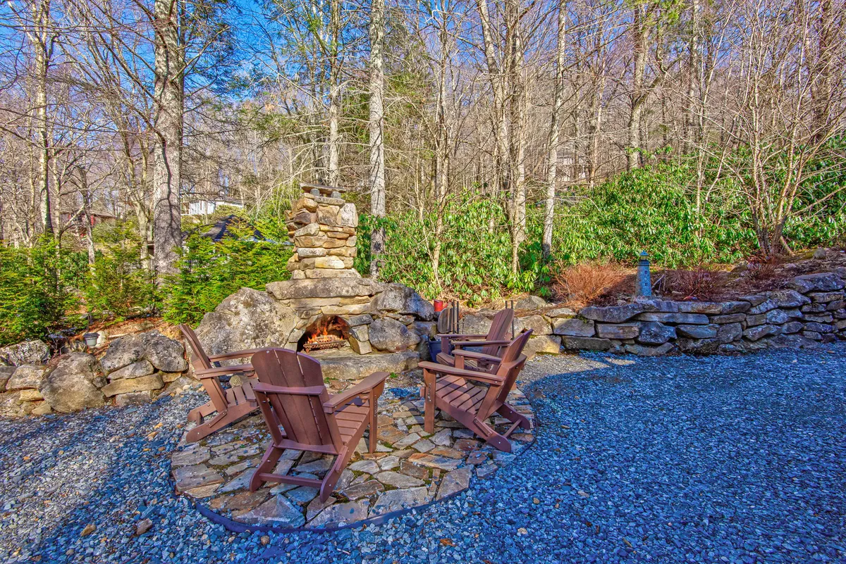 A charming outdoor stone fireplace is surrounded by Adirondack chairs, set amidst tall trees and a gravel path. The ambiance invites intimate gatherings and cozy moments by the fire, with a clear view of the surrounding nature.