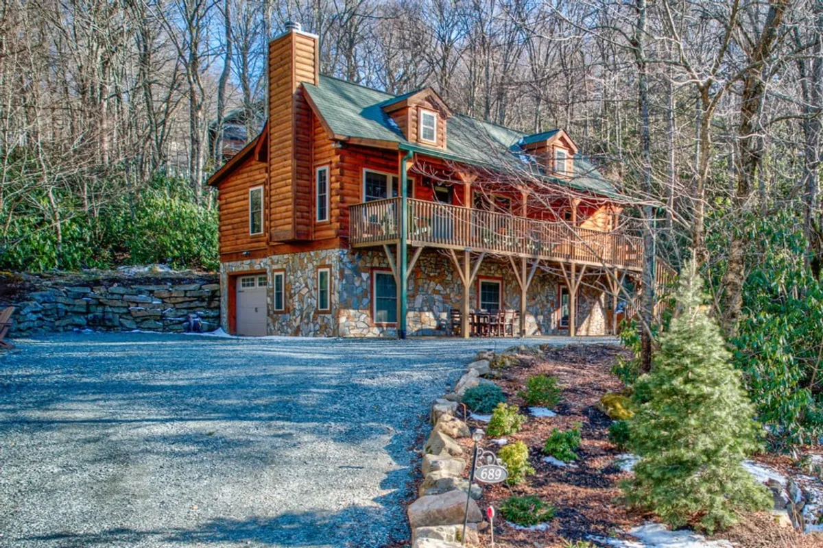 A charming log cabin with a welcoming wooden balcony and elegant stone facade, set amidst lush greenery. The spacious gravel driveway leads up to the home, while the balcony and outdoor areas offer perfect spots for relaxing in nature or gathering around the fire pit.