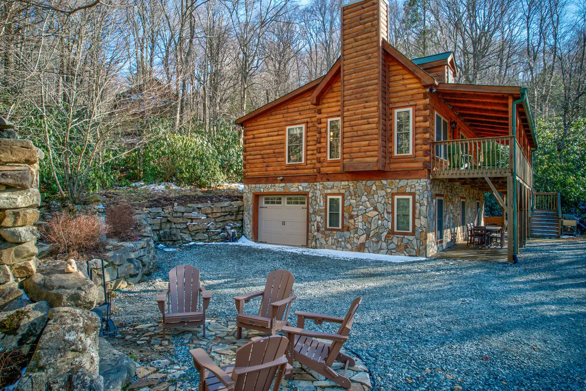 This exterior image displays the rustic log cabin structure of Squirrel’s Leap, featuring a stone foundation, Adirondack chairs arranged around a fire pit, and a beautiful wooded setting. The cozy front porch and abundant trees reflect the inviting atmosphere of summer mountain escapes.