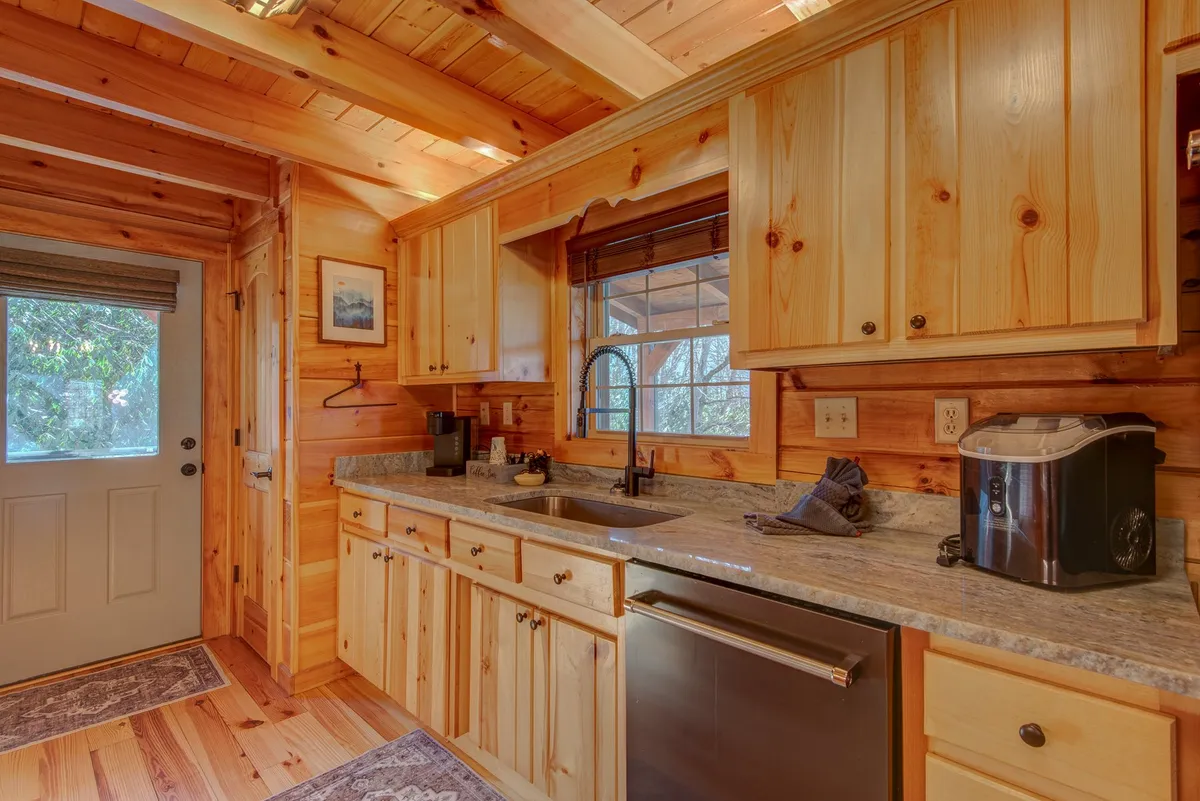 This image shows a warmly lit kitchen featuring a granite countertop, wooden cabinets, a kitchen sink with a window letting in natural light, and a door leading outside. The scene exudes the feeling of a central gathering space where family meals and conversations unfold.