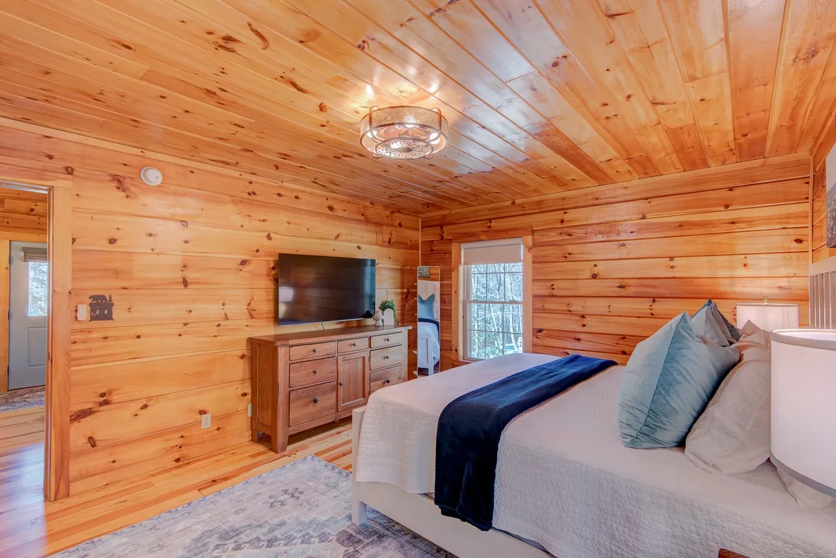 A serene bedroom with wooden walls, a neatly made bed adorned with plush pillows, a dresser, and a TV. Natural light streams into the room, creating a restful atmosphere ideal for relaxation and undisturbed sleep.